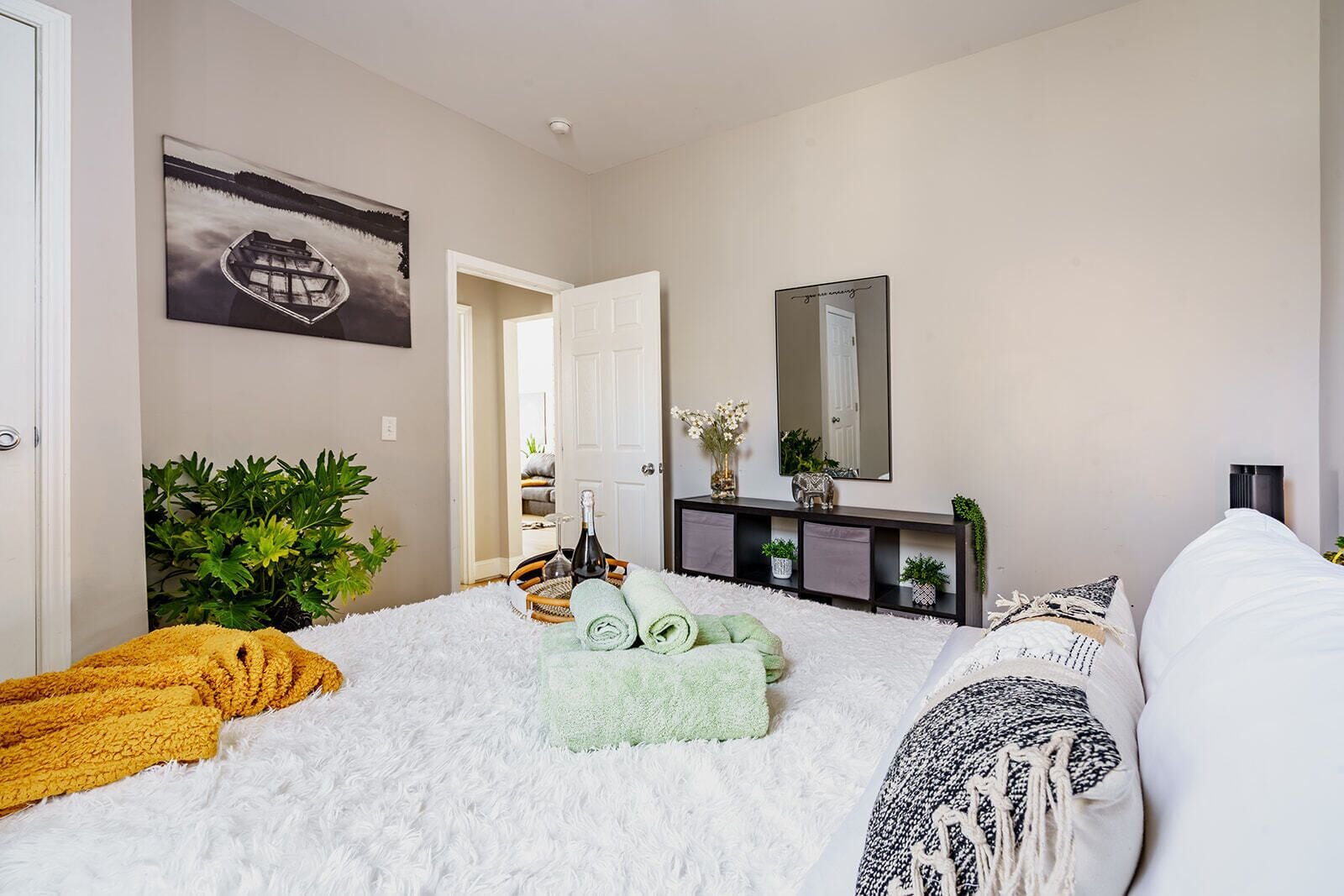 Een slaapkamer met een zacht, wit tapijt, een bed met witte en gemêleerde kussens, een nachtkastje met een fles en glazen, een console tafel met planten, en een schilderij aan de muur.