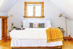 A bedroom with a large bed, a wooden headboard, a window with blinds, a bedside lamp, and a nightstand with a bottle and glasses. - Jungle House Boutique Lofts – Downtown Columbus & Short North (Columbus)