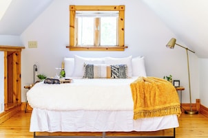 Un dormitorio con una cama grande, cabecera de madera, ventana con persianas, lámpara de noche y mesita de noche con una botella y vasos.
