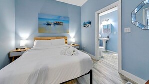 A bedroom with a bed, two bedside lamps, a painting of boats, a bathroom with a sink and mirror, and a wooden floor. - Jungle House Boutique Lofts – Downtown Columbus & Short North (Columbus)