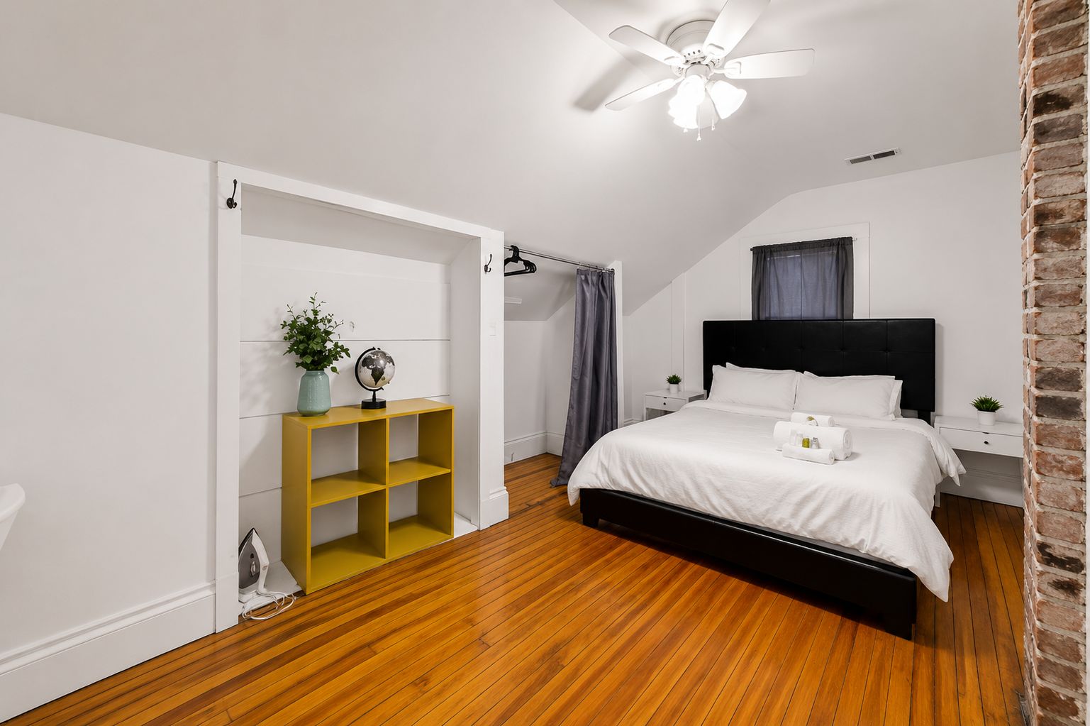 A bedroom with a bed, a shelf with a globe and plant, a ceiling fan, and a brick wall.