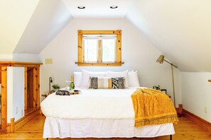 A bedroom with a sloped ceiling, a large bed with white bedding, a wooden floor, a window with blinds, and a bedside lamp.