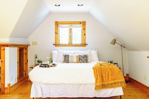 Un dormitorio con techo inclinado, una cama grande con ropa de cama blanca, suelo de madera, una ventana con persianas y una lámpara de noche.