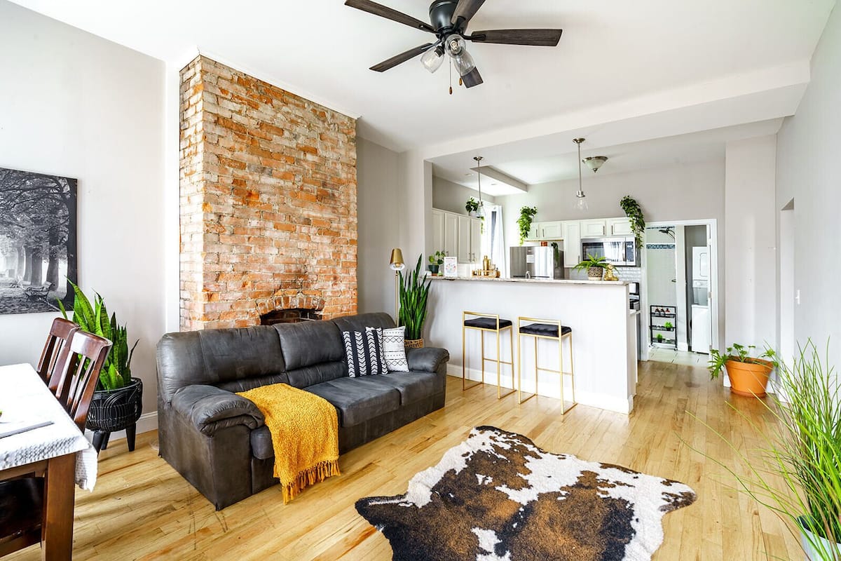 A modern living room with a brick wall, a leather sofa, a cowhide rug, and a kitchen in the background.