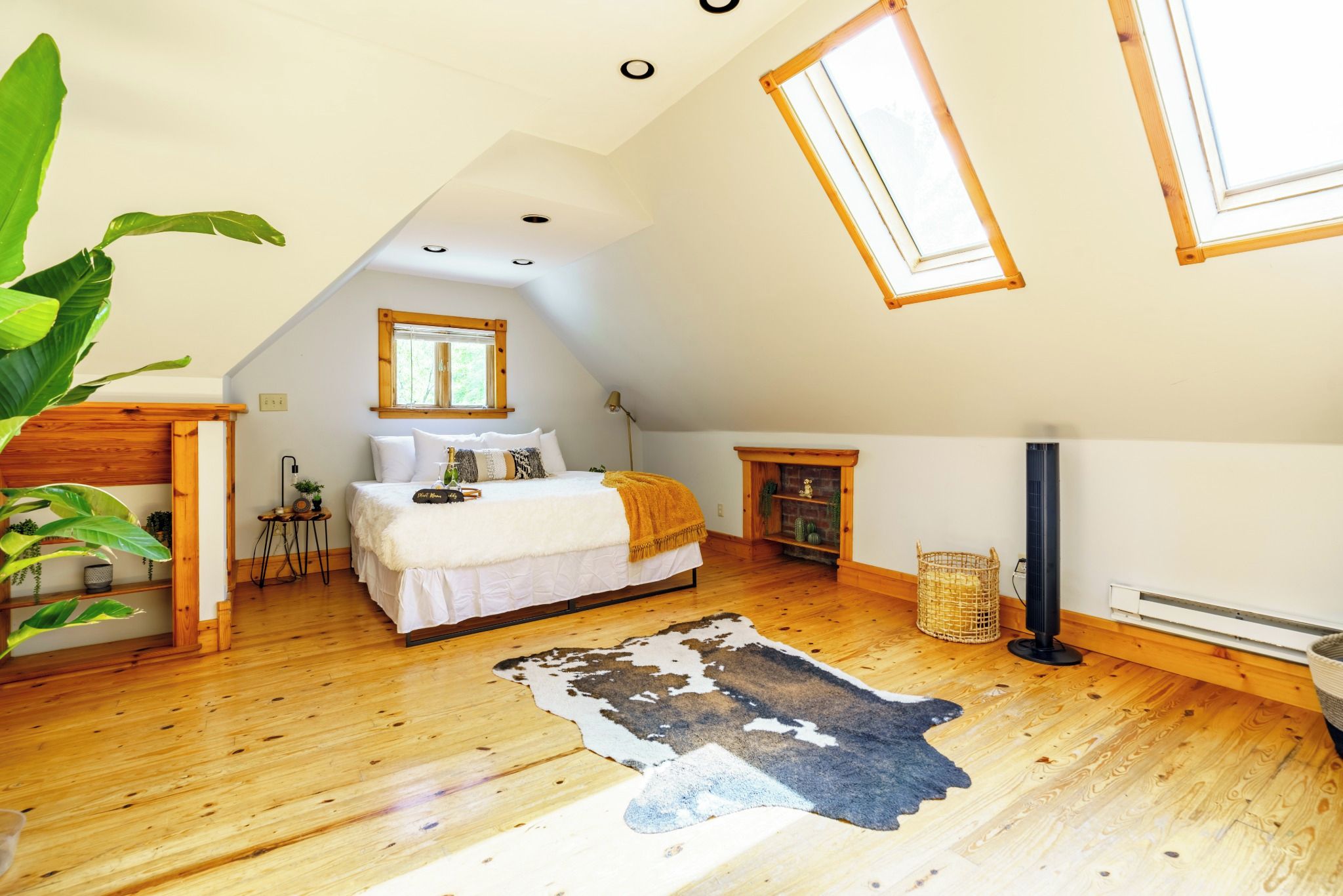 A bedroom with a sloped ceiling, wooden flooring, a bed with white bedding, a cowhide rug, a small fireplace, and a window with a wooden frame.