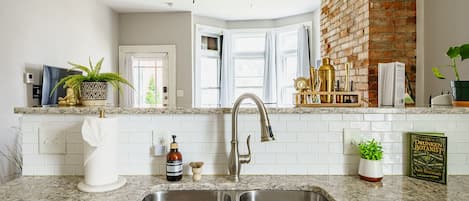 A kitchen with a double sink, a ceiling fan, a brick wall, and a window with curtains.