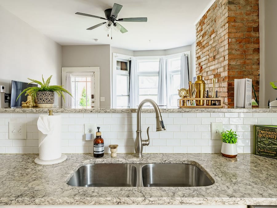 A kitchen with a double sink, a ceiling fan, a brick wall, and a window with curtains.