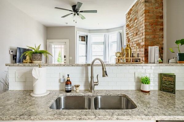 A kitchen with a double sink, a ceiling fan, a brick wall, and a window with curtains.