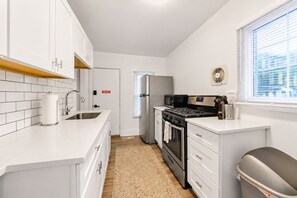 A compact kitchen with white cabinetry, a stainless steel sink, a built-in oven, and a window with blinds. - Jungle House Boutique Lofts – Downtown Columbus & Short North (Columbus)