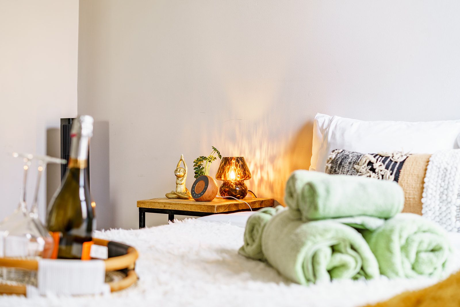 A neatly made bed with green towels, a bedside table with a lamp, a bottle, and a small statue.