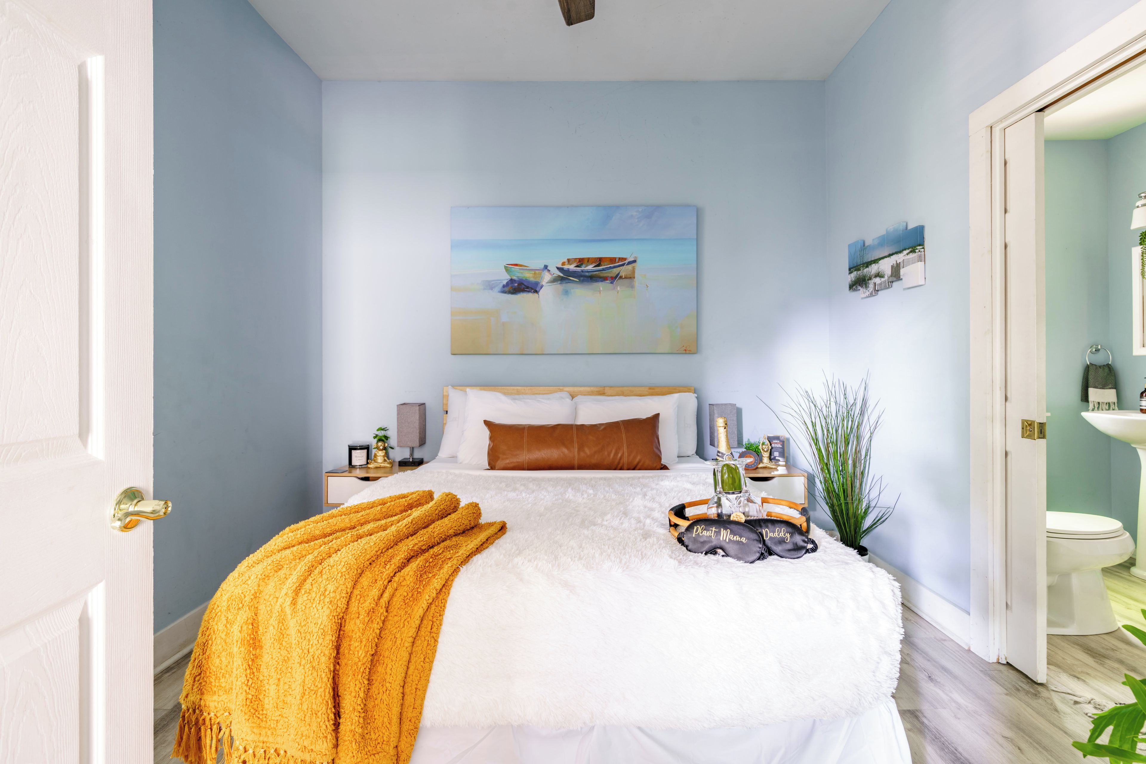 Une chambre à coucher avec un lit, un tableau représentant des bateaux, une table de chevet avec une lampe et une petite table de nuit avec une bouteille.