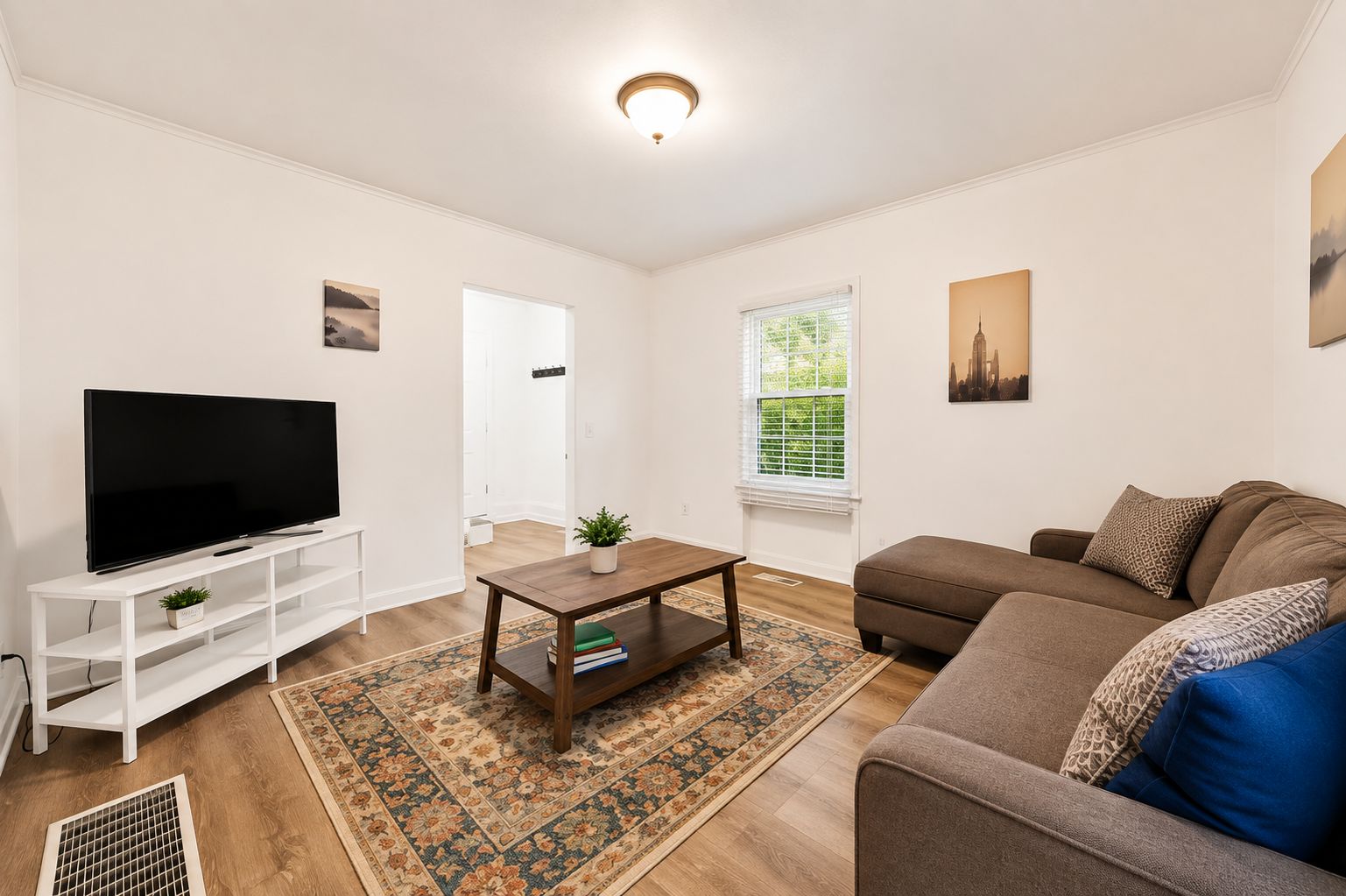 Ein Wohnzimmer mit einem Flachbildfernseher auf einem weißen Schrank, einem braunen Sofa mit gemusterten Kissen, einem Holztisch und einem Teppich mit Muster.