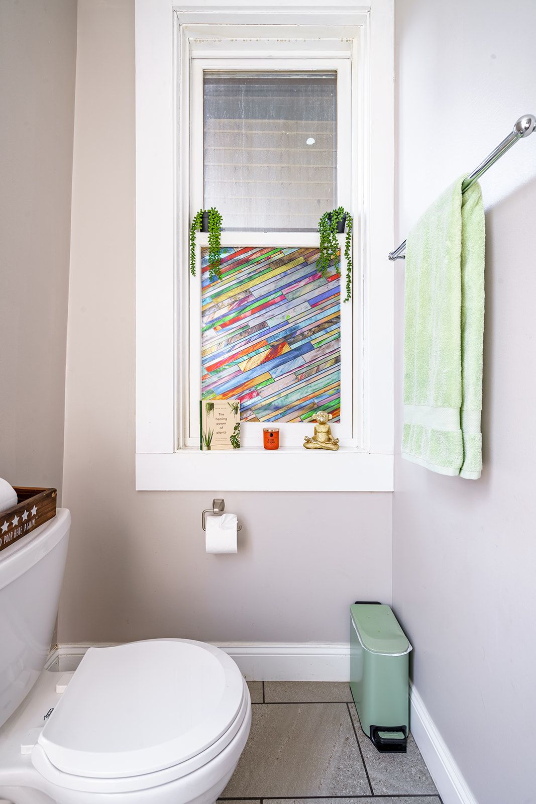 Een badkamer met een wit toilet, een raam met een kleurrijk houten slalendesign, een groene handdoek en een kleine prullenbak.