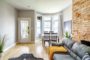 A living room with a grey sofa, a dining table with chairs, a large window with curtains, and a brick wall.