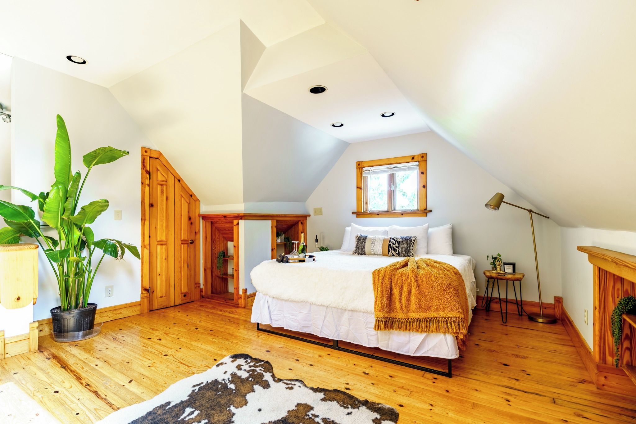 A bedroom with a sloped ceiling, a bed with white bedding, a wooden floor, a large potted plant, and a small table with a lamp.