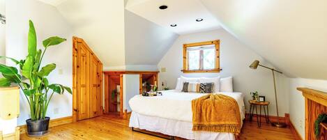 A bedroom with a sloped ceiling, a bed with white bedding, a wooden floor, a large potted plant, and a small table with a lamp.