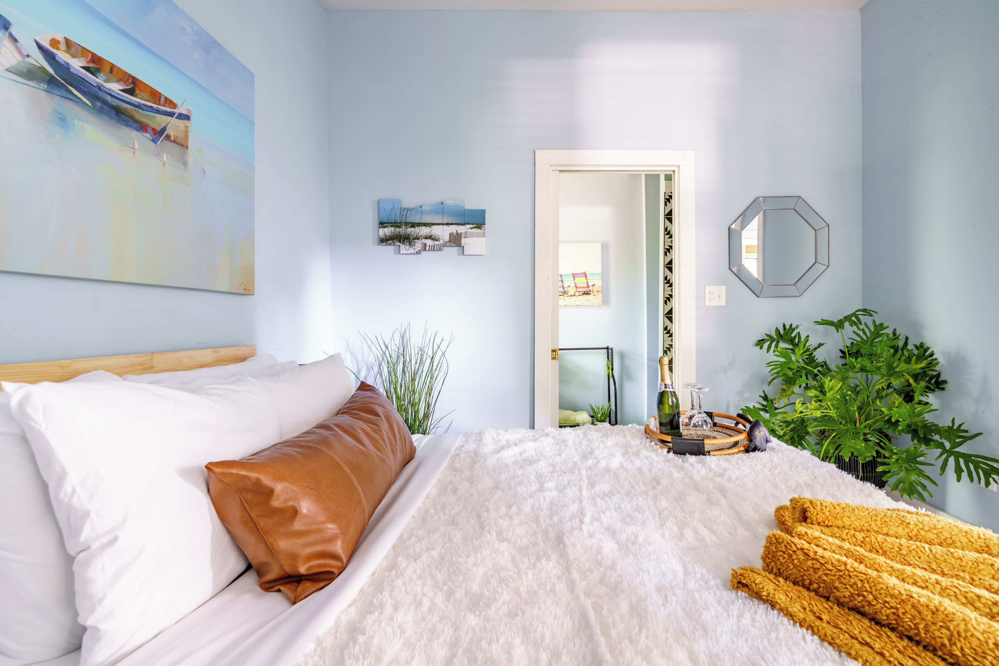 A bedroom with a bed, a painting, a mirror, and a plant.