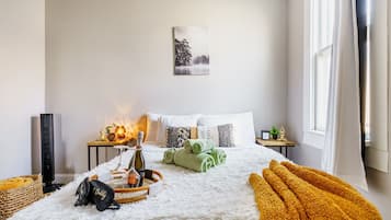 Een slaapkamer met een bed, kussens, een nachtkastje met een lamp, een raam en een schilderij aan de muur.