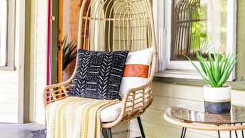 A wicker chair with a cushion and a throw blanket, positioned next to a window with a view of greenery.