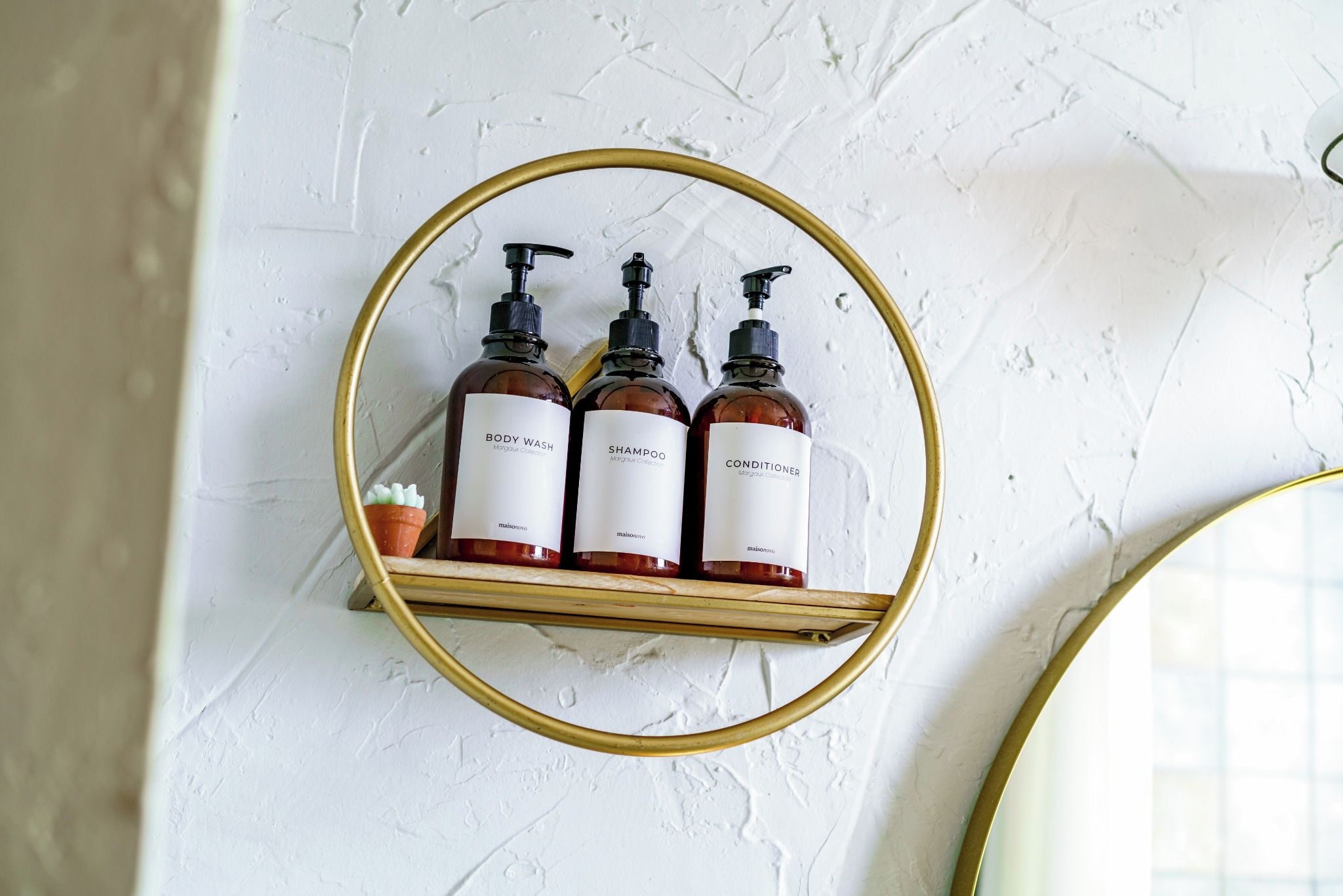 A white textured wall with a gold-framed mirror shelf holding three bottles of body wash, shampoo, and conditioner.