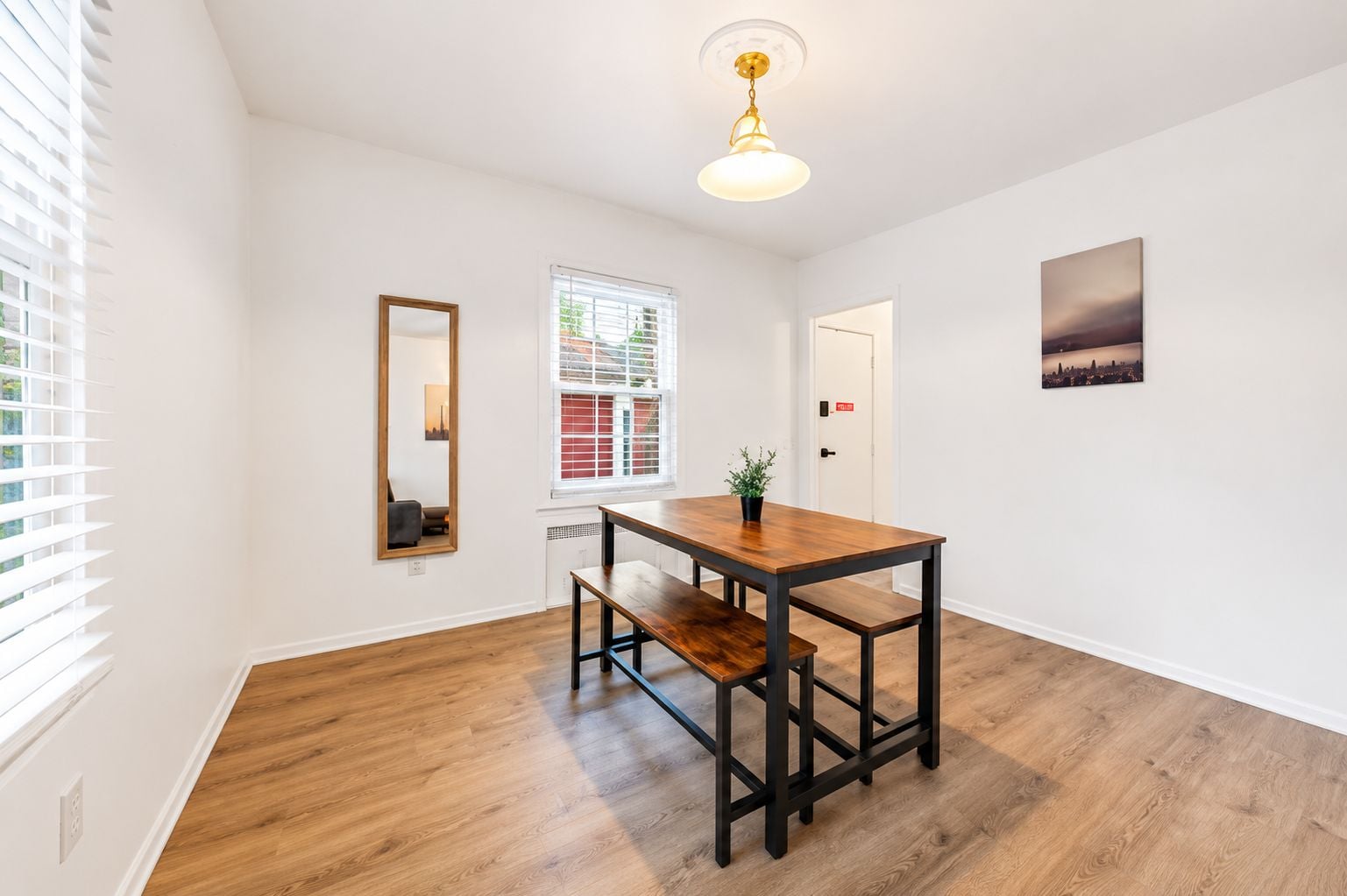 Una habitación con una mesa de comedor de madera, una banca, un espejo, una ventana con persianas, una puerta y una pintura de un paisaje urbano.