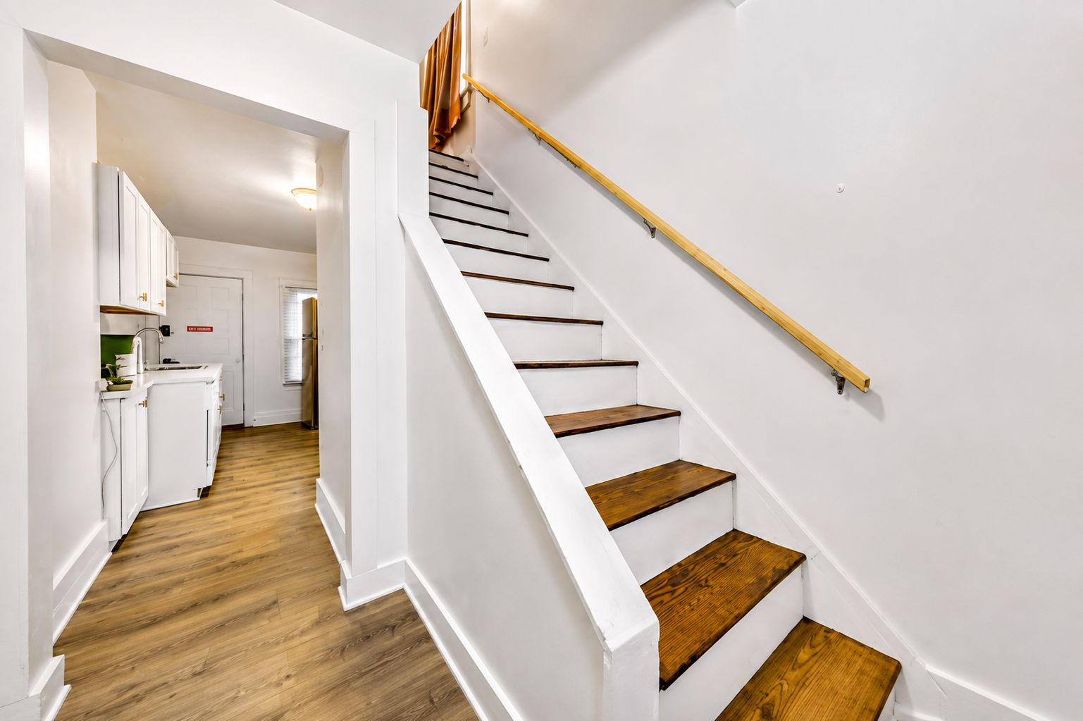 A modern interior with a staircase leading to a second floor, a kitchen area with white cabinetry, and wooden flooring.