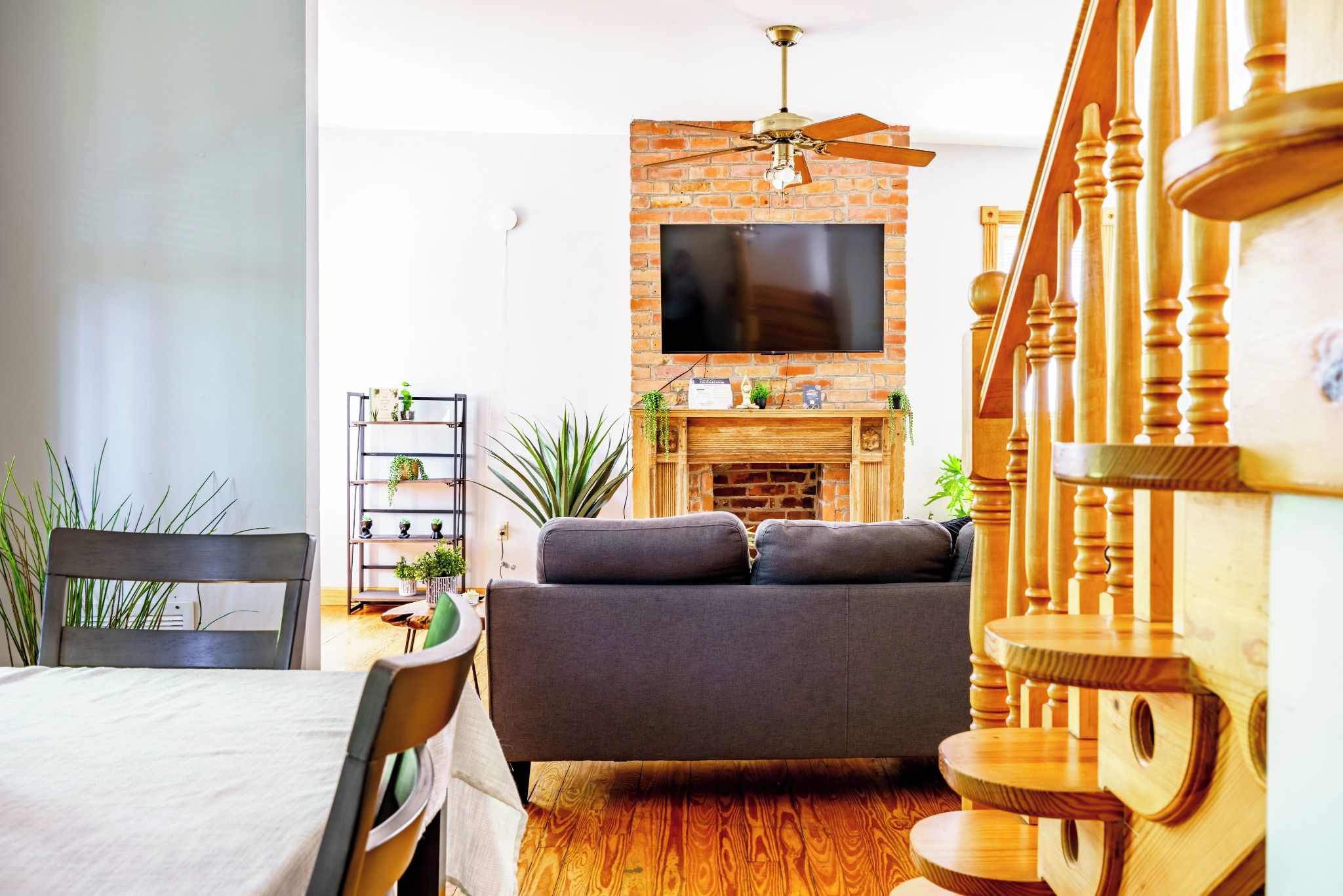 A living room with a sofa, a television mounted on a brick wall, a wooden staircase, and a dining area with a table and chairs.