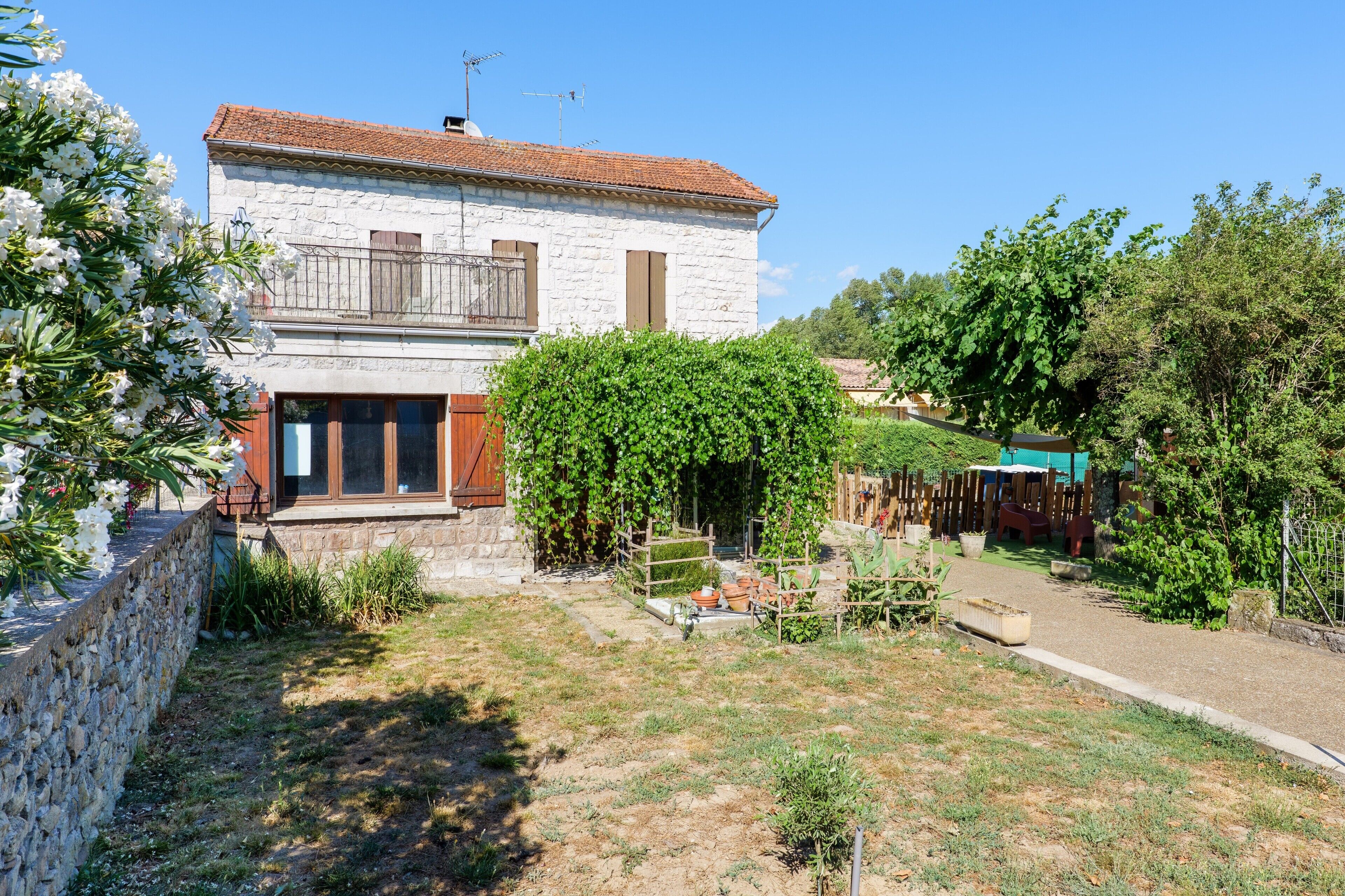 Un bâtiment en pierre avec un toit en tuiles, un balcon et un jardin agrémenté de plantes et d’une clôture.