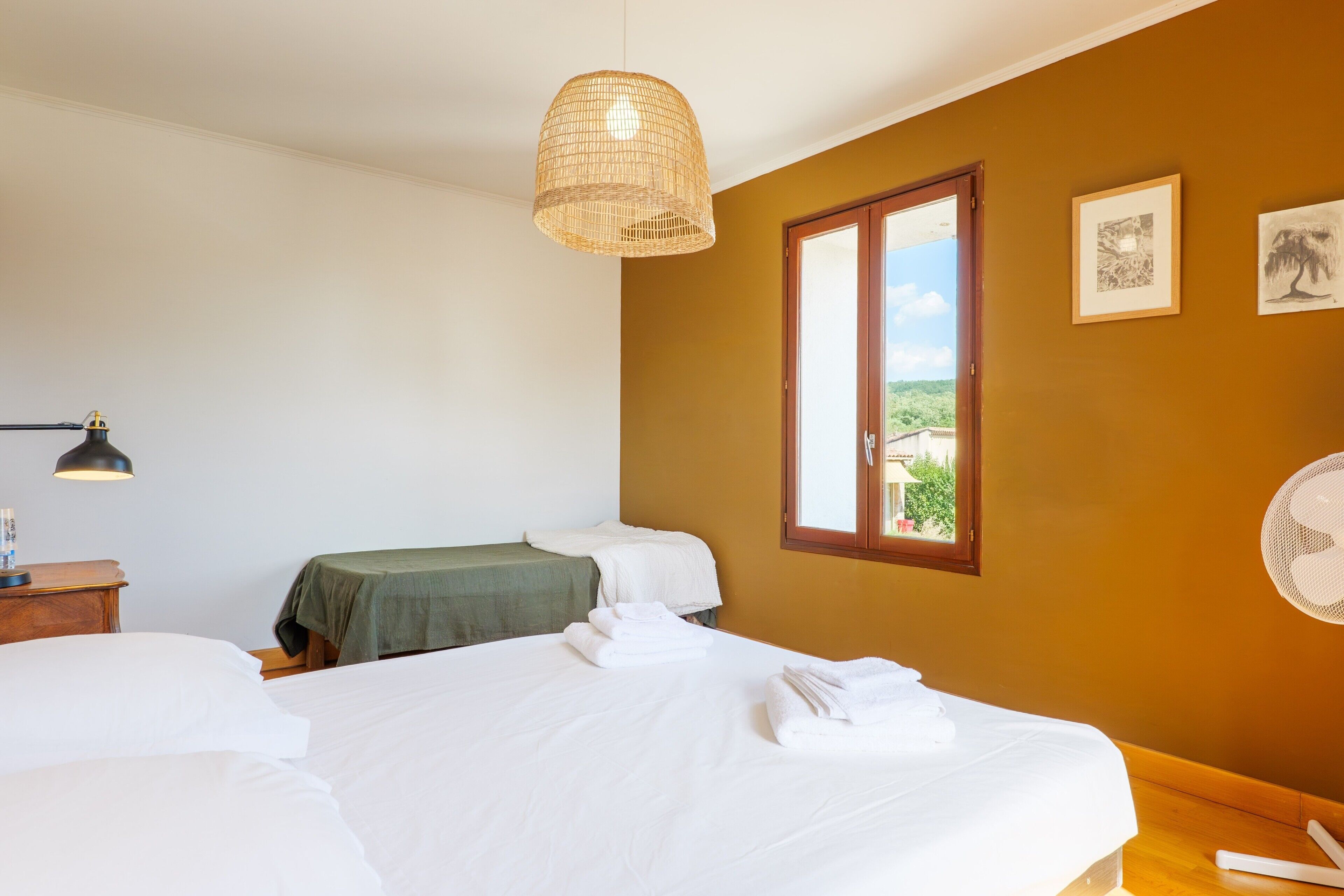 A hotel room with a bed, a bedside table, a lamp, a window, and a wall-mounted fan.