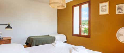 A hotel room with a bed, a bedside table, a lamp, a window, and a wall-mounted fan.