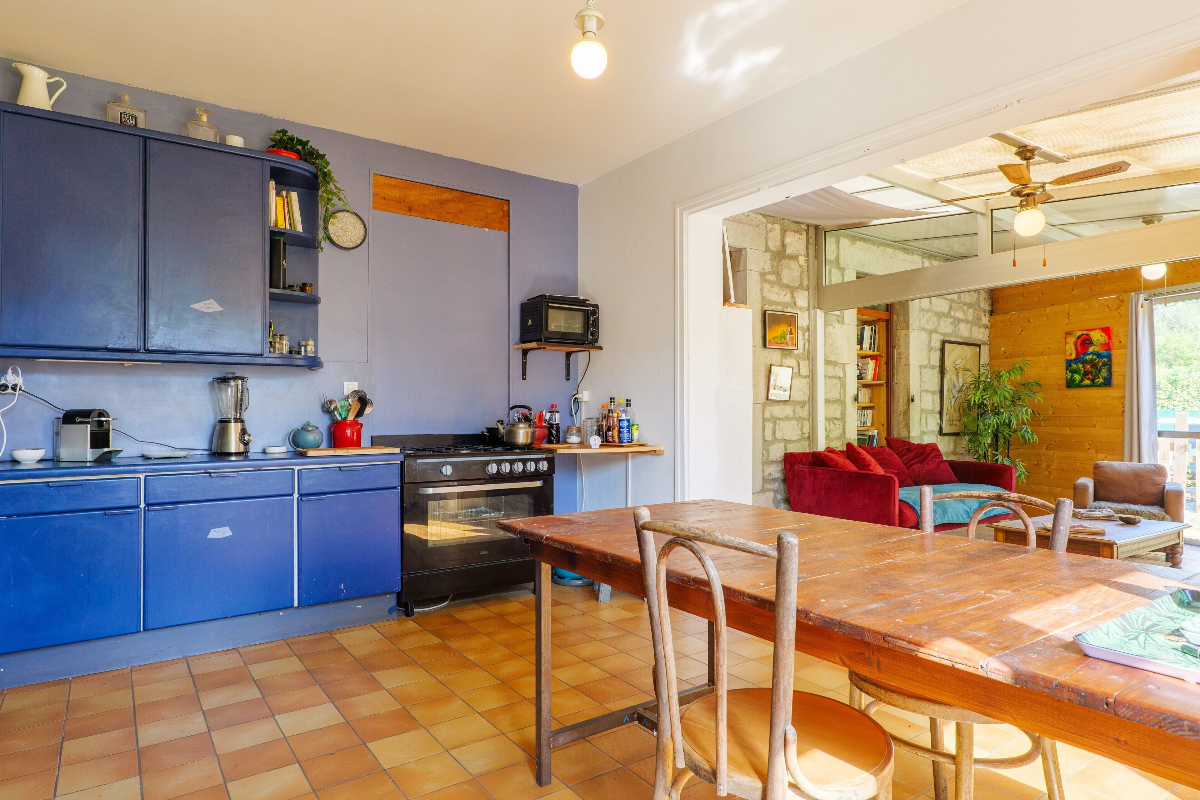Une cuisine avec des meubles de couleur bleue, une table à manger en bois et un canapé rouge dans l’espace salon.