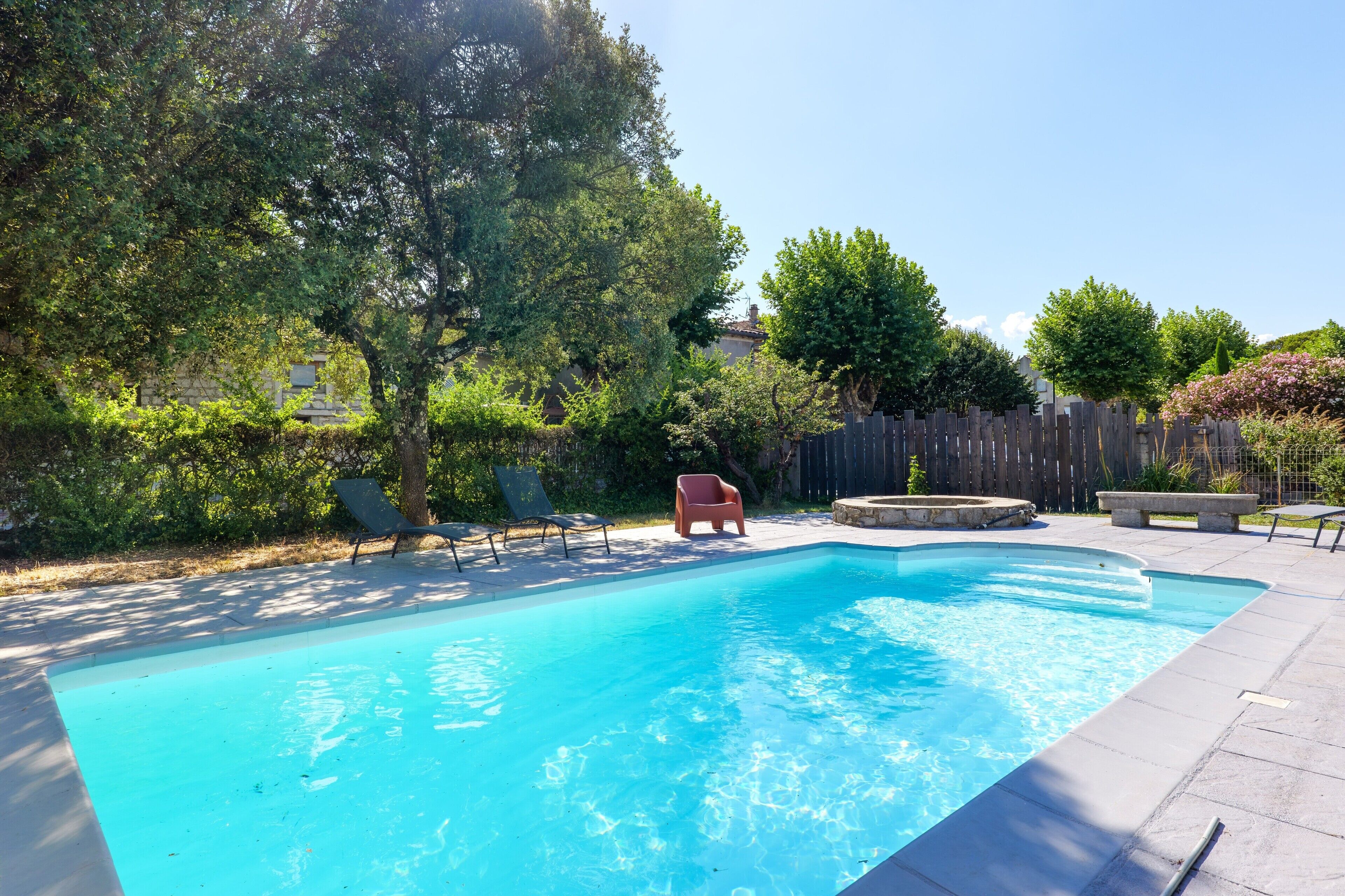A rectangular swimming pool with a wooden deck surrounding it, a wooden fence, and a few trees.