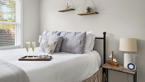 Un lit bien fait, sur lequel on a posé un plateau avec des coupes à champagne et une maquette de bateau, une lampe de chevet et deux plantes suspendues.