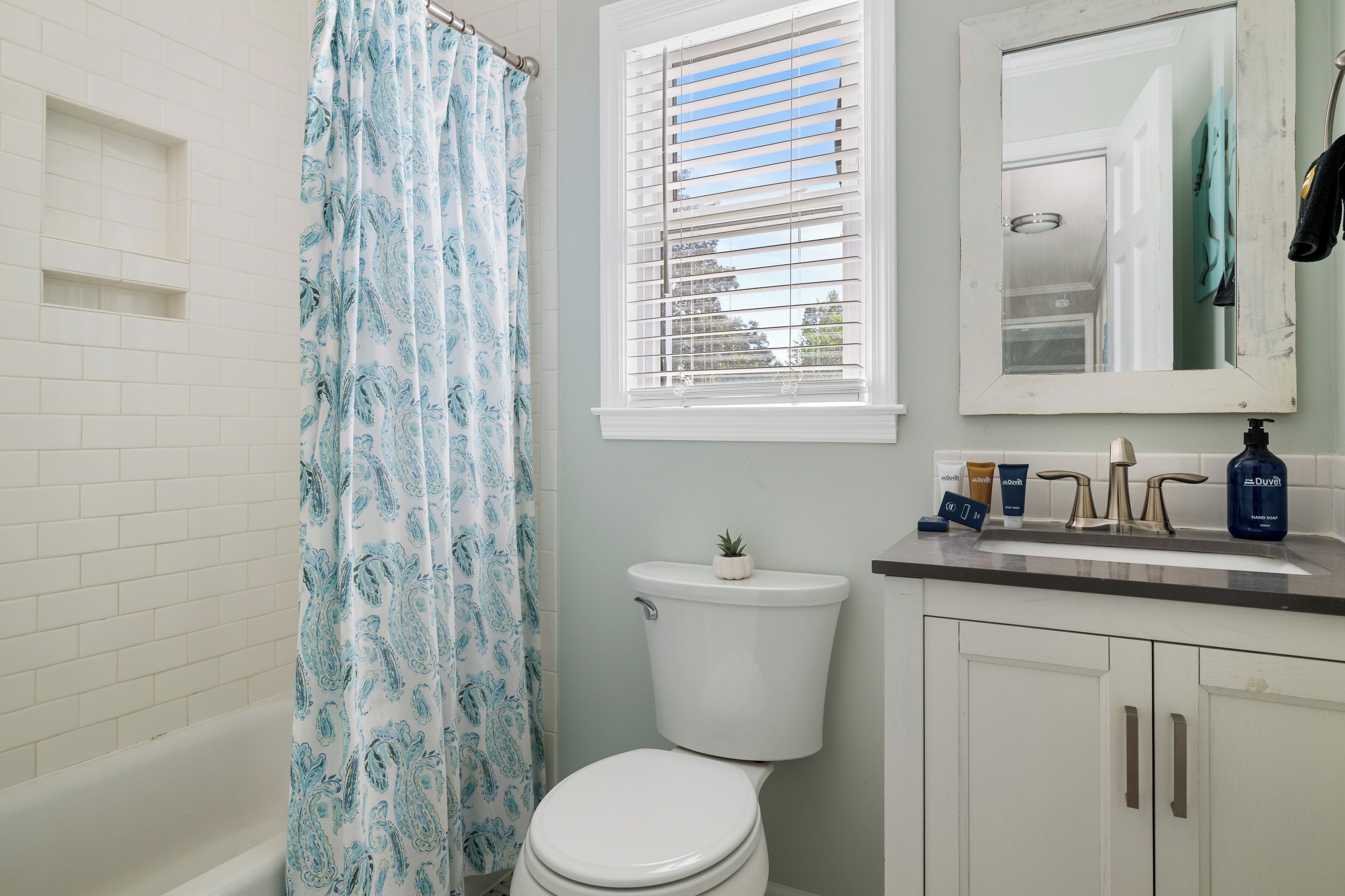 Une salle de bain comprenant des toilettes, un rideau de douche, une fenêtre avec des stores, un lavabo avec des articles de toilette et un miroir.