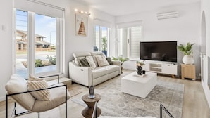 A modern living room with a sofa, a coffee table, a TV, and large windows. - The Coastal Haven 1BR (Shoalwater)