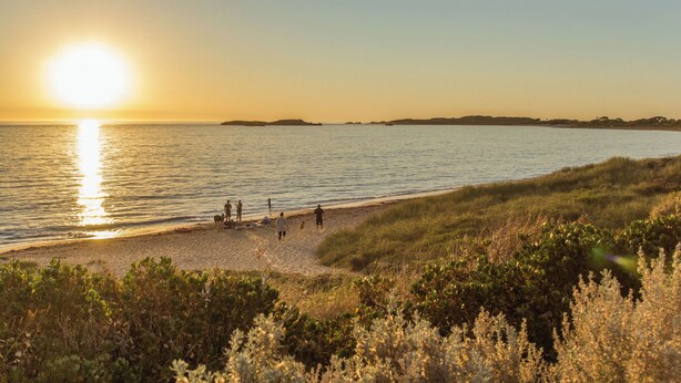 A beach with people walking and sitting on the sand, and a sunset over the water. - The Coastal Haven 1BR (Shoalwater)