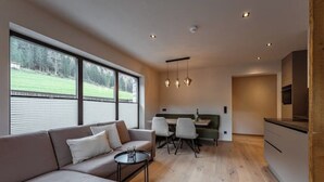 A modern living room with a sofa, dining table, and chairs. There is a kitchen area with a stove and cabinets. Large windows provide a view of the outdoors.
