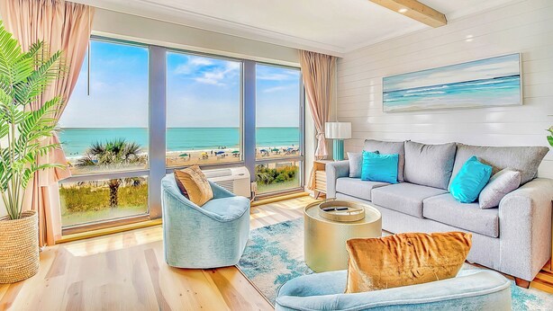 A spacious living room with large windows overlooking a beach, a grey sofa with blue cushions, and a potted plant. - Modern Awesome Jacuzzi Romance Condo w 2 bathrooms (Myrtle Beach)