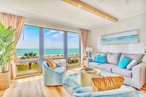 A spacious living room with large windows overlooking a beach, a grey sofa with blue cushions, and a potted plant. - Modern Awesome Jacuzzi Romance Condo w 2 bathrooms (Myrtle Beach)