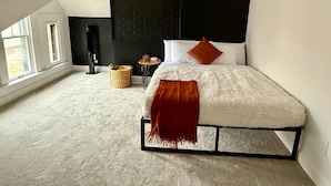 Un dormitorio con techo inclinado, una cama con ropa de cama blanca y un plaid rojo, una mesita auxiliar pequeña, una cesta tejida y un cabecero negro.