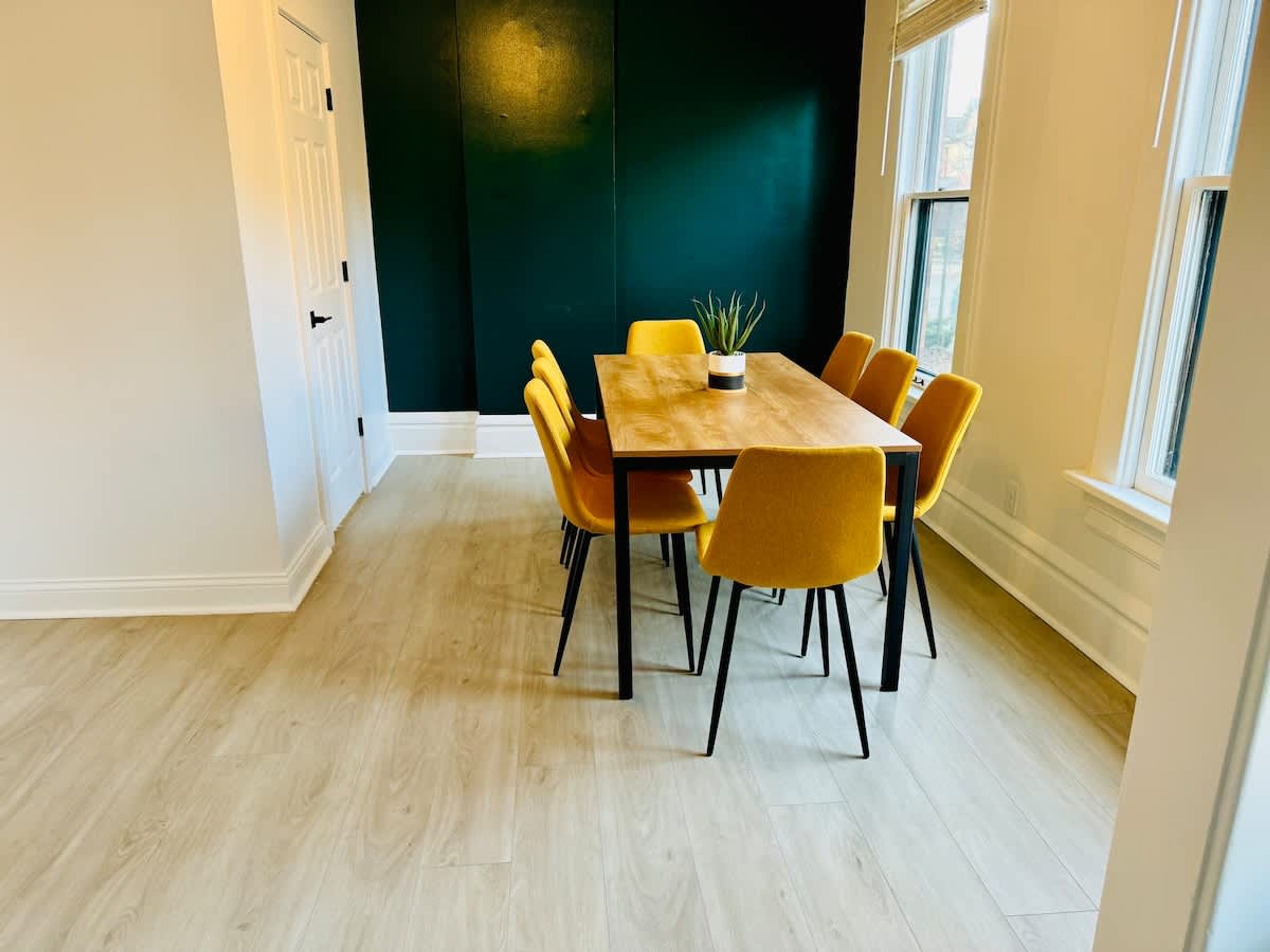A dining area with a wooden table and yellow chairs, a potted plant, and a dark green accent wall.