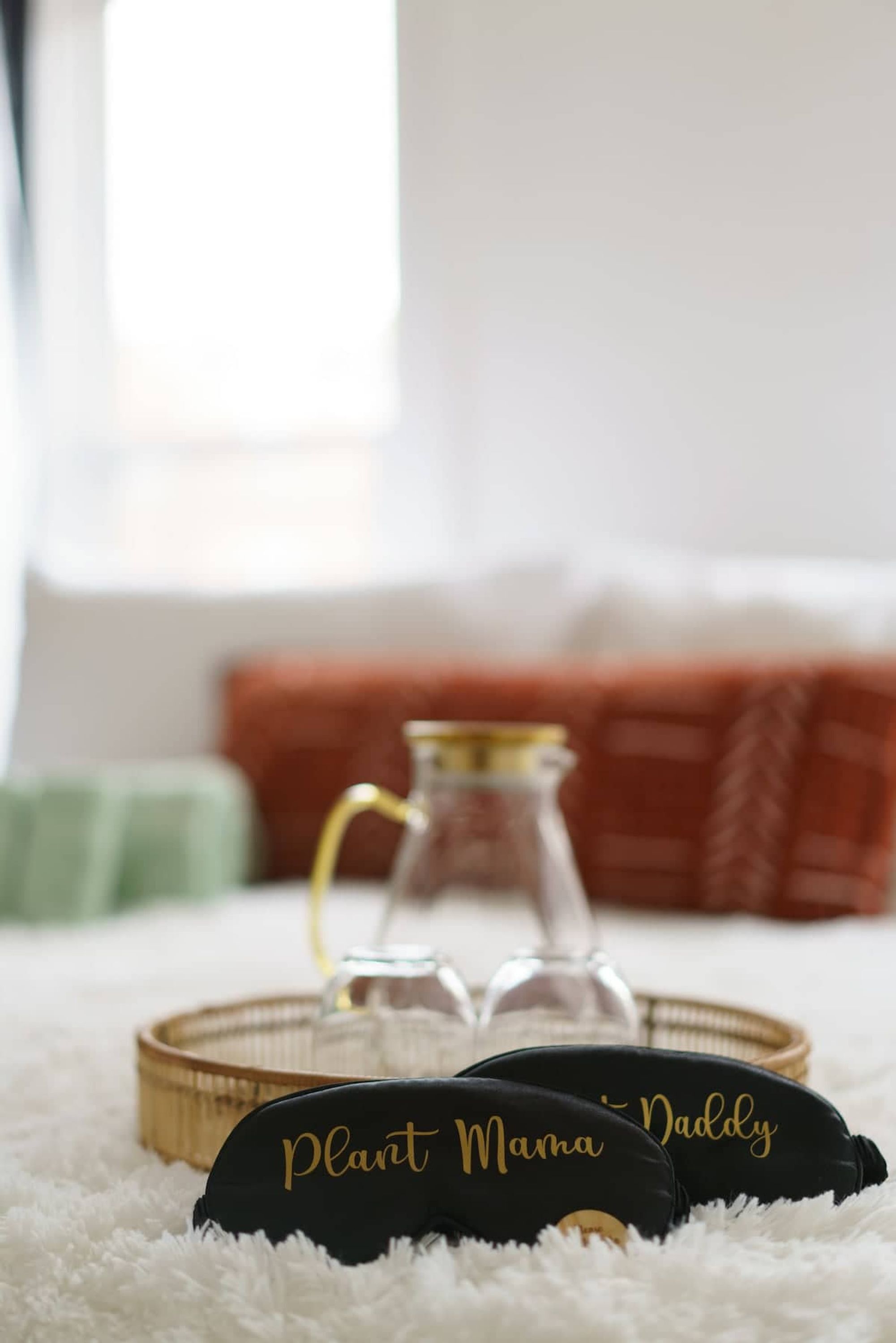 Ein gemütliches Zimmer mit einem weißen, flauschigen Teppich, einem schwarzen Kissen mit der Aufschrift 'Plant Mama Daddy' und einem klaren Glasbehälter sowie einer gewebten Ablagefläche.