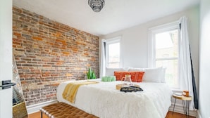 A bedroom with a brick wall, a white bed with a fluffy white cover, a brown bench, and a small table with a cup. - Prime Location Top Rated Clean Parking Free (Columbus)