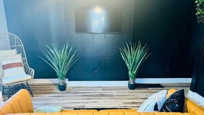 Un salón moderno con un sofá amarillo, dos plantas en macetas, un sillón de mimbre y una pared oscura con una ventana.