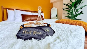 Una cama con almohadas blancas, una almohada negra con el texto "Plant Mama" y un decantador de vidrio, además de una manta amarilla.