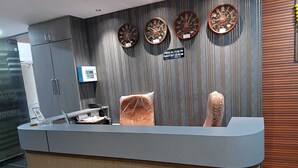 A reception desk with a check-in/out sign, a digital display, and a wall with clocks. - Hotel R Town (Vadodara)