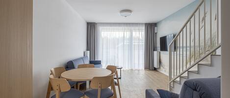 Una sala de estar moderna con una mesa redonda de madera, sillas azules, un sofá gris y una escalera con barandilla.