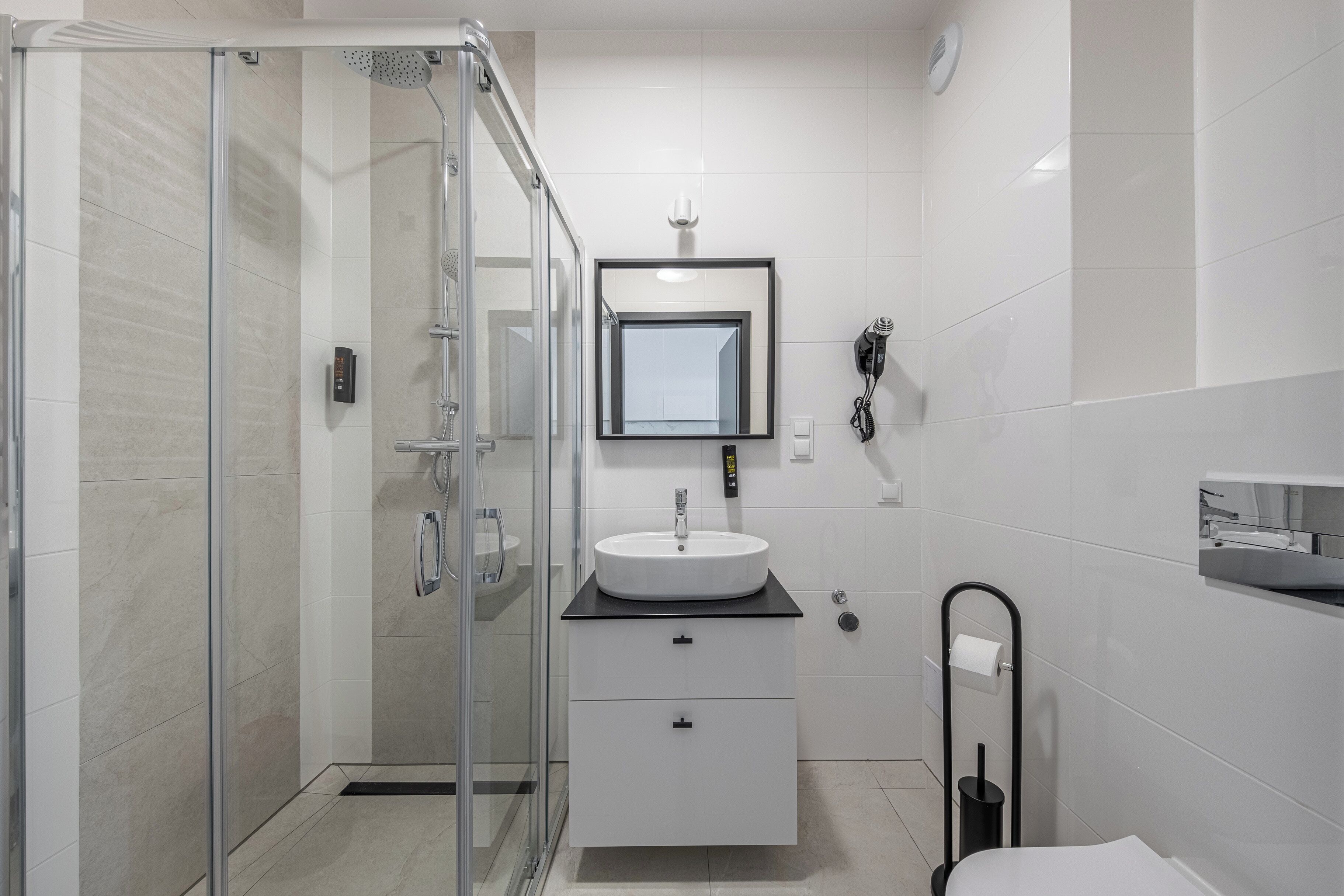 Un bagno moderno con box doccia in vetro, lavandino bianco su piano nero, specchio sopra il lavandino e water con portarotolo nero.
