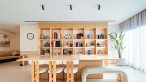 A modern living room with a wooden bookshelf, a dining table with chairs, a bench, and a large window with curtains. - Hamilton House Studio: Rooftop Pool & Gym Access (Dubai)