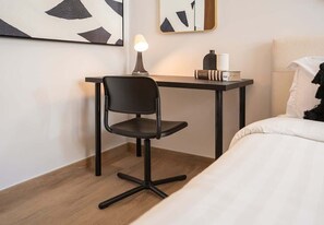 A modern hotel room with a wooden floor, a black office chair, a desk with a lamp, a mirror, and a bed with white bedding.